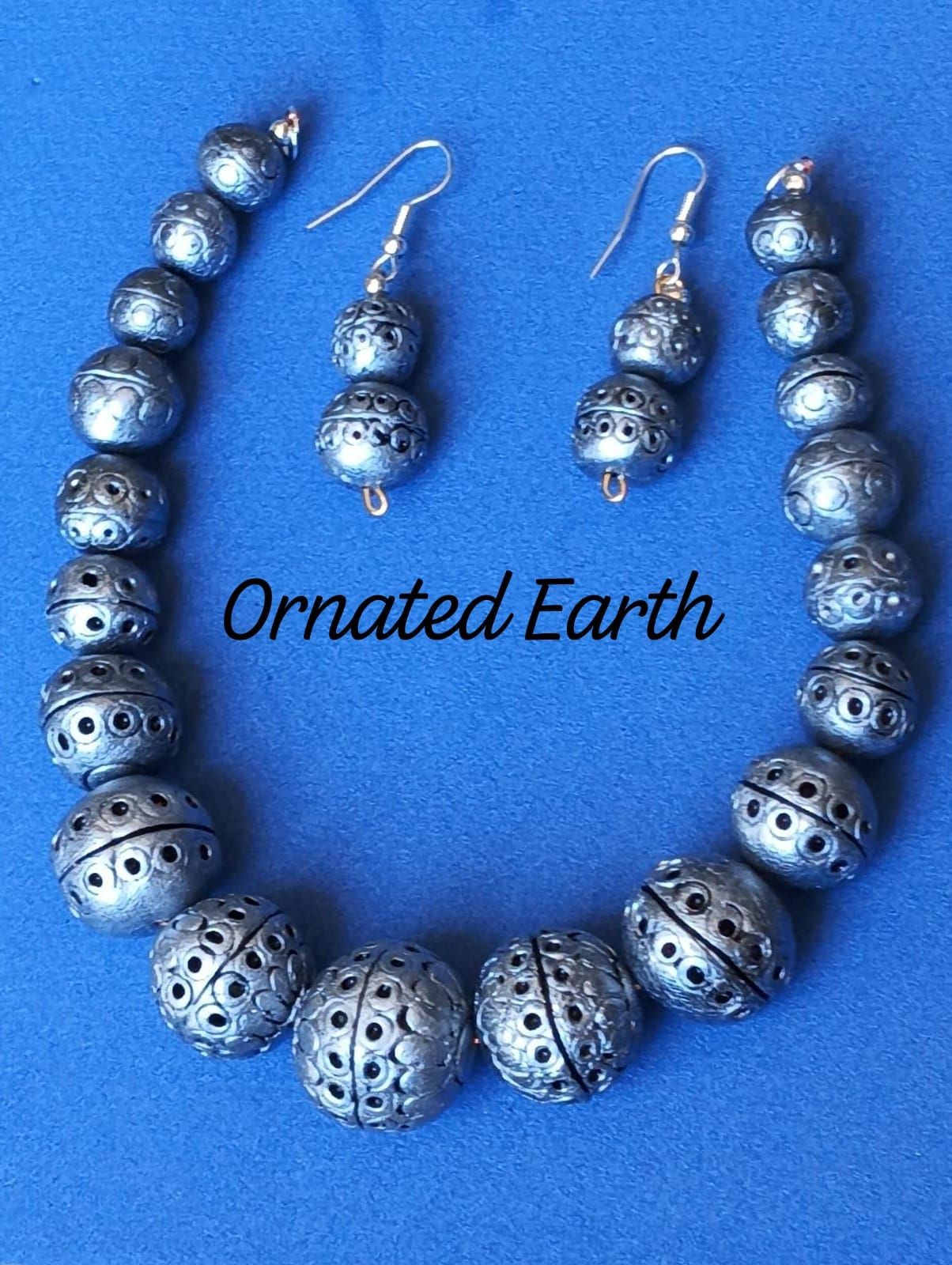 Silver beaded necklace and earrings terracotta clay set on blue fabric, labeled 'Ornated Earth.'